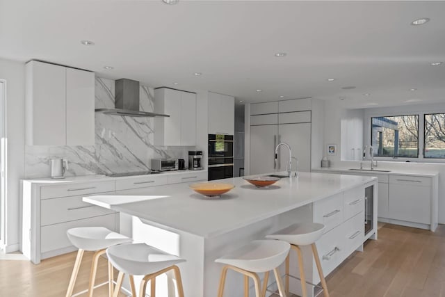 kitchen featuring light countertops, white cabinets, a center island with sink, and wall chimney range hood