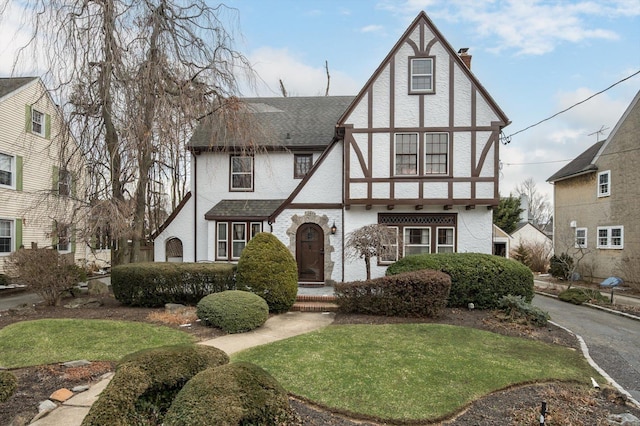 tudor house featuring a front yard, a shingled roof, a chimney, and stucco siding