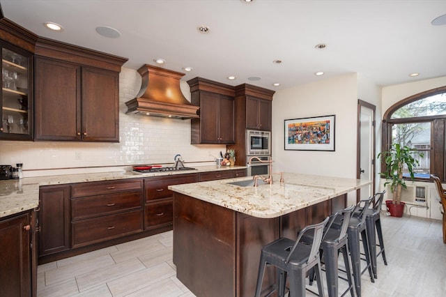 kitchen featuring light stone counters, stainless steel appliances, custom range hood, glass insert cabinets, and a kitchen island with sink