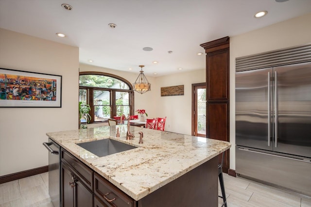 kitchen featuring stainless steel appliances, light stone countertops, a sink, dark brown cabinets, and an island with sink