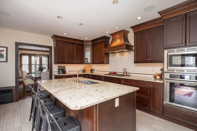 kitchen featuring stainless steel appliances, glass insert cabinets, a sink, an island with sink, and premium range hood