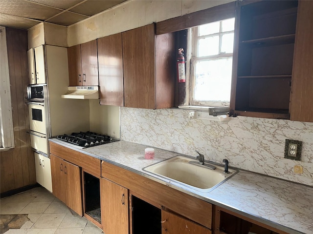 kitchen with brown cabinets, black gas cooktop, light countertops, a sink, and under cabinet range hood