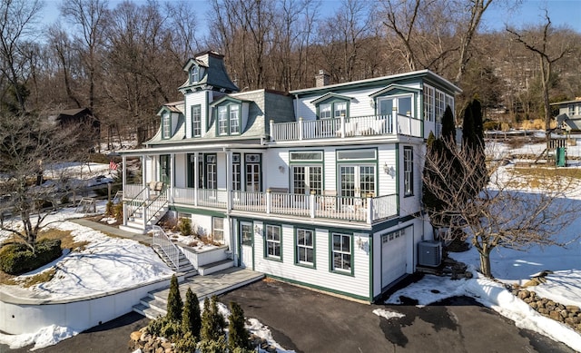 snow covered rear of property featuring mansard roof, central AC unit, aphalt driveway, a balcony, and stairway