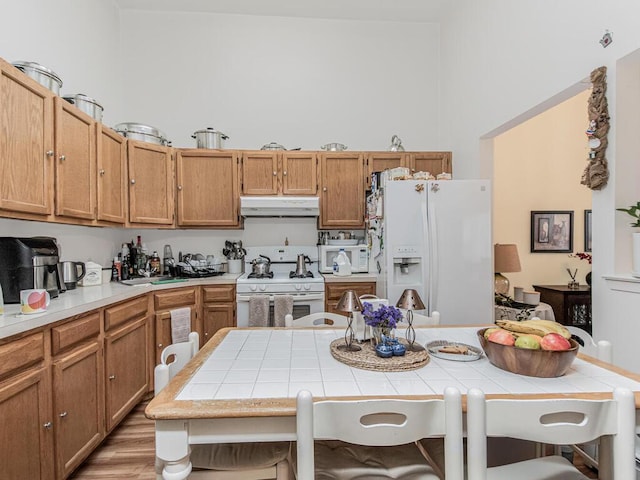 kitchen featuring white appliances, light wood finished floors, tile counters, a high ceiling, and under cabinet range hood