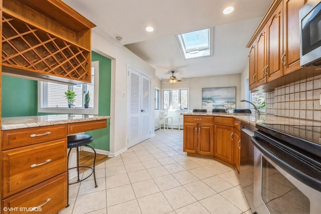 kitchen featuring a skylight, decorative backsplash, appliances with stainless steel finishes, brown cabinetry, and light stone countertops