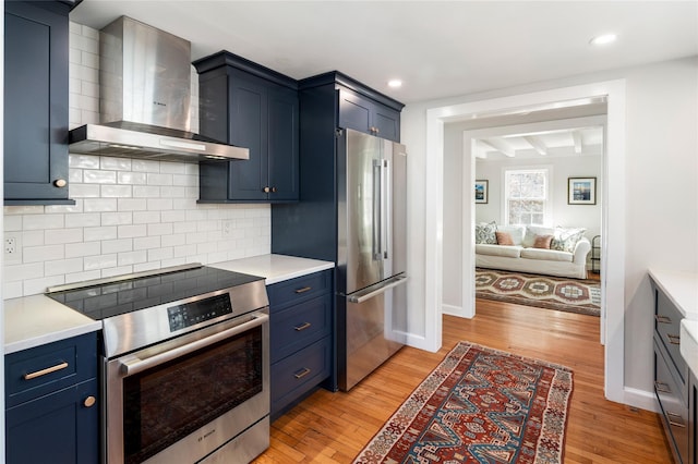 kitchen with wall chimney range hood, blue cabinetry, appliances with stainless steel finishes, and light wood-style floors