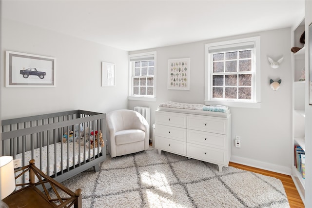 bedroom with radiator, a nursery area, baseboards, and wood finished floors