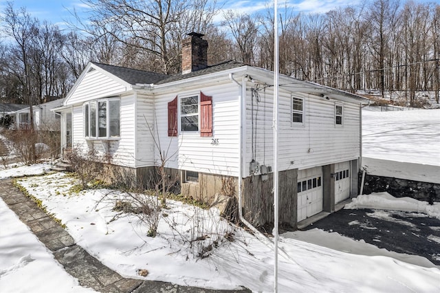 snow covered property featuring driveway, a chimney, and an attached garage