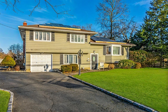 split level home with aphalt driveway, a chimney, solar panels, a front yard, and a garage