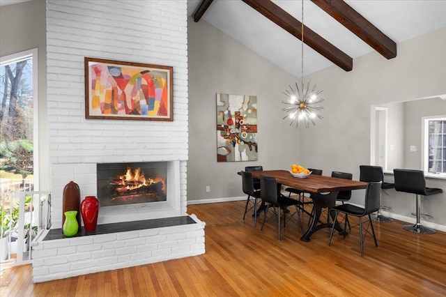 dining space with a brick fireplace, a notable chandelier, wood finished floors, and beamed ceiling