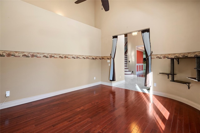 empty room with wood-type flooring, a towering ceiling, a ceiling fan, baseboards, and stairs