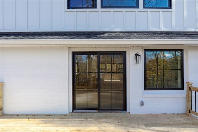 property entrance featuring board and batten siding, roof with shingles, brick siding, and a wooden deck