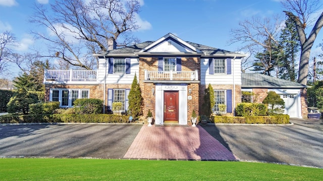 colonial house with a garage, a balcony, a chimney, a front lawn, and brick siding