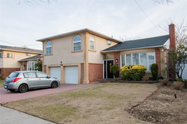 split level home with decorative driveway, brick siding, a chimney, stucco siding, and an attached garage