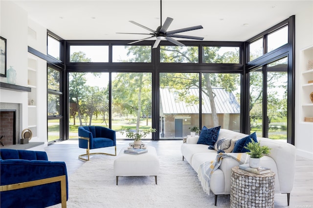 living area with built in features, a fireplace with raised hearth, a ceiling fan, expansive windows, and light wood-type flooring