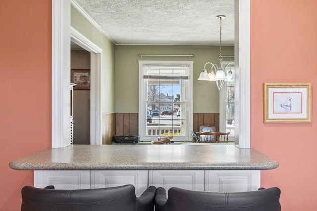 kitchen with a wainscoted wall, crown molding, an inviting chandelier, white cabinets, and a textured ceiling