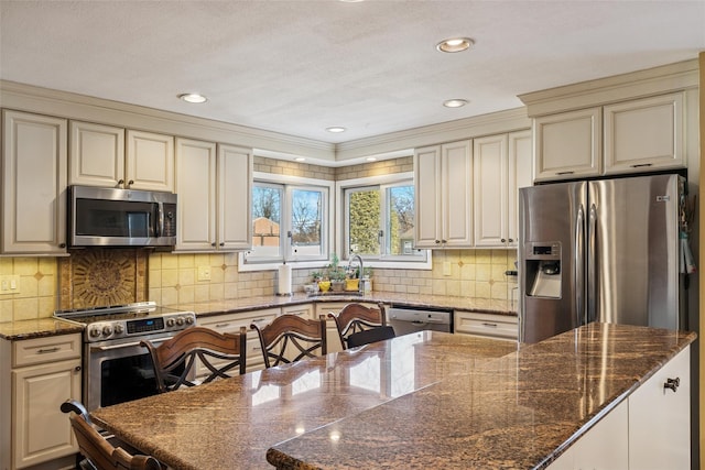 kitchen featuring cream cabinetry, stainless steel appliances, backsplash, a sink, and dark stone counters