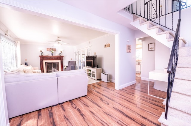 living room with a ceiling fan, stairway, crown molding, light wood-type flooring, and a fireplace