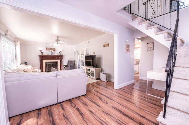 living room featuring a ceiling fan, wood finished floors, stairs, crown molding, and a fireplace