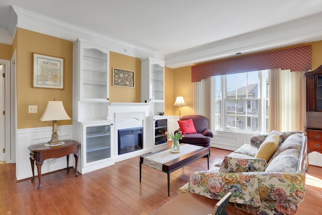 living room with wood-type flooring, a glass covered fireplace, crown molding, and wainscoting