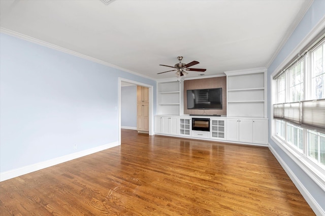 unfurnished living room featuring built in shelves, wood finished floors, a ceiling fan, baseboards, and crown molding