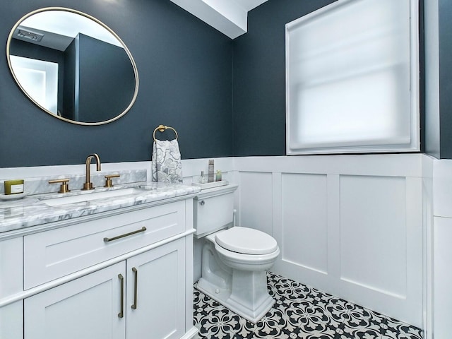 half bathroom featuring wainscoting, toilet, tile patterned floors, vanity, and a decorative wall