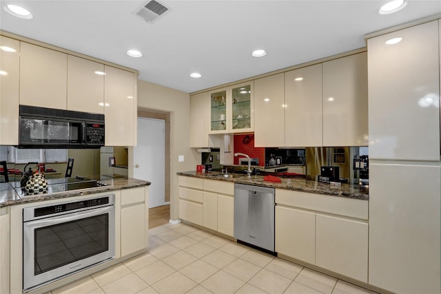 kitchen with black appliances, cream cabinets, glass insert cabinets, and dark stone countertops