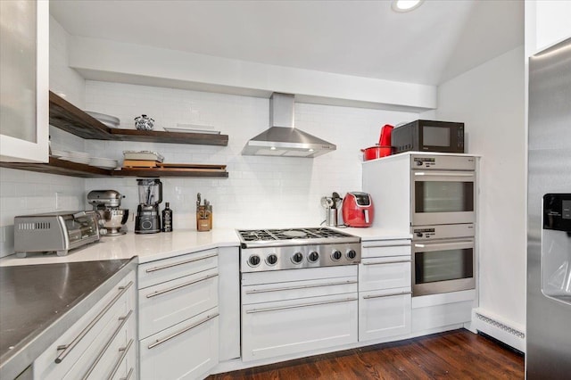 kitchen featuring tasteful backsplash, dark wood-style floors, appliances with stainless steel finishes, baseboard heating, and wall chimney range hood