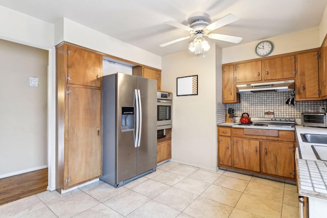 kitchen with brown cabinets, decorative backsplash, appliances with stainless steel finishes, ceiling fan, and under cabinet range hood