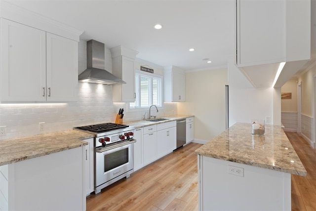 kitchen featuring light stone counters, stainless steel appliances, white cabinetry, a sink, and wall chimney range hood