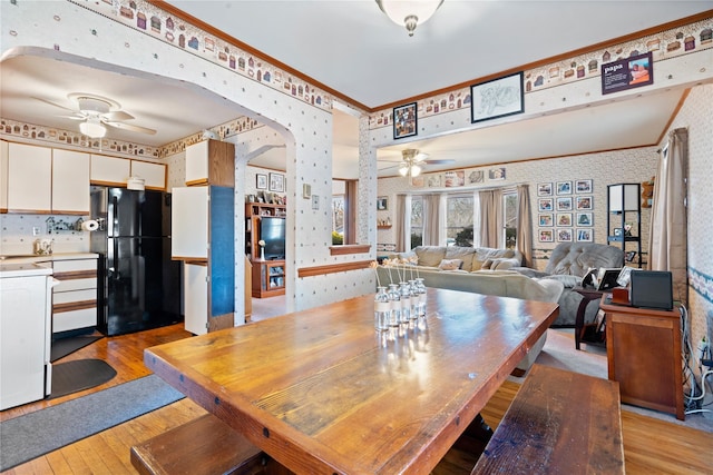 dining room featuring arched walkways, crown molding, light wood-style floors, ceiling fan, and wallpapered walls