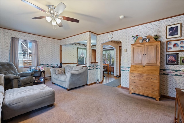 carpeted living area featuring arched walkways, a ceiling fan, baseboards, wallpapered walls, and crown molding