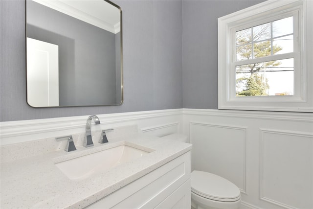 half bathroom featuring toilet, a decorative wall, a wainscoted wall, vanity, and ornamental molding