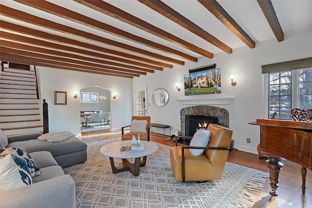 living area featuring a warm lit fireplace, baseboards, stairway, wood finished floors, and beam ceiling