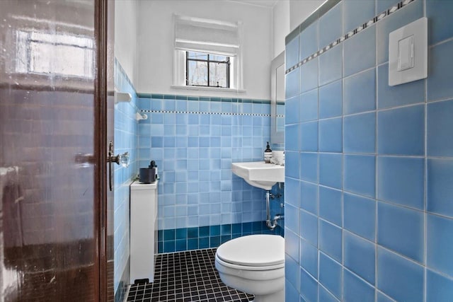 half bath with tile patterned flooring, toilet, a wainscoted wall, a sink, and tile walls