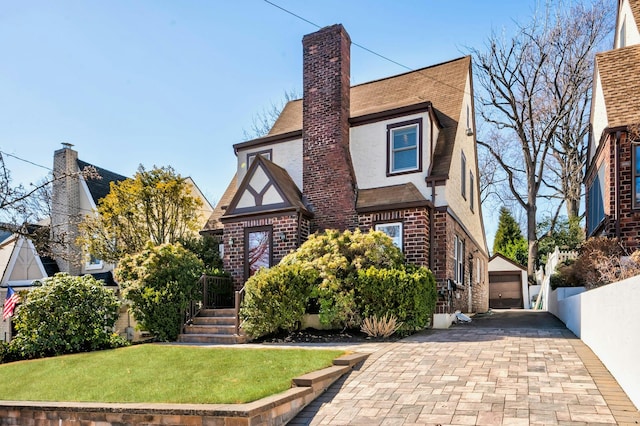 tudor-style house with an outbuilding, brick siding, a chimney, stucco siding, and a front yard