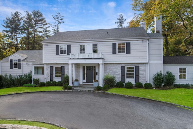 view of front of house with a shingled roof, a balcony, stone siding, a chimney, and a front yard