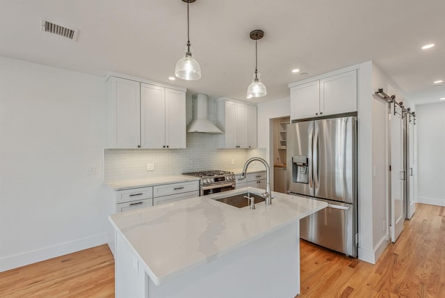 kitchen featuring a barn door, appliances with stainless steel finishes, white cabinets, a kitchen island with sink, and wall chimney range hood