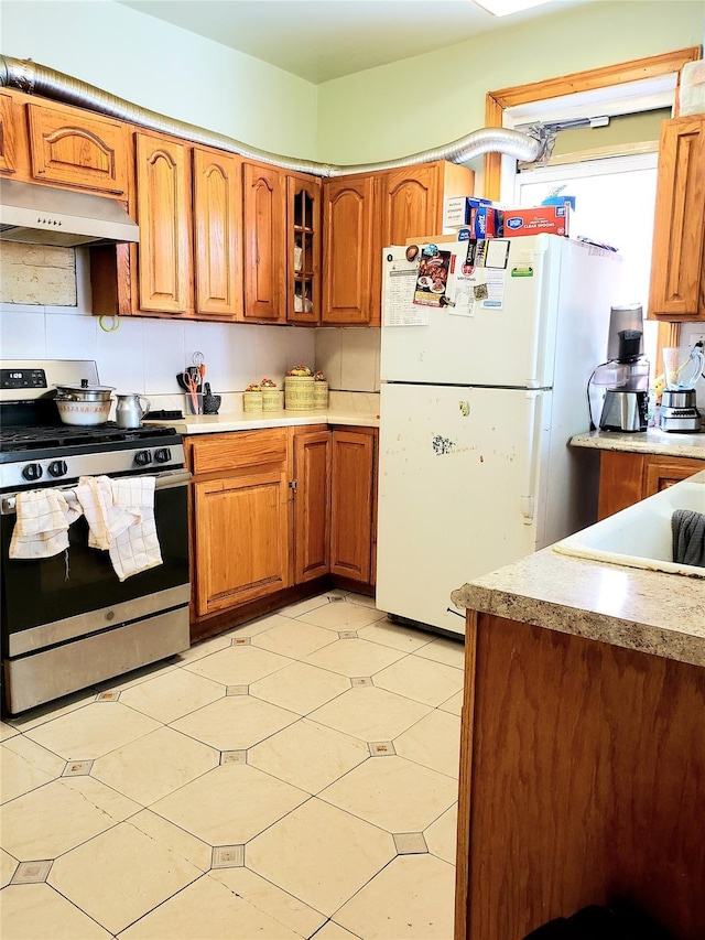 kitchen featuring stainless steel gas stove, under cabinet range hood, brown cabinets, and freestanding refrigerator