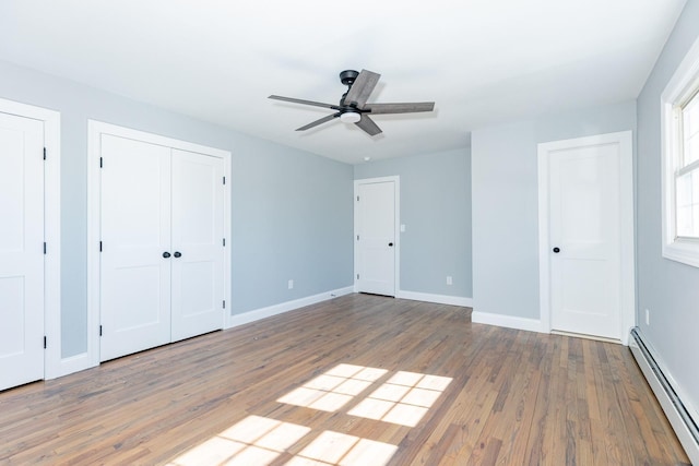 unfurnished bedroom featuring two closets, a baseboard heating unit, a ceiling fan, wood finished floors, and baseboards