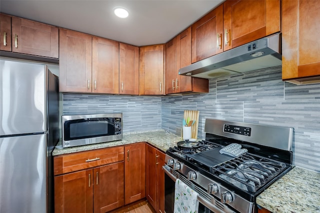 kitchen featuring backsplash, under cabinet range hood, light stone counters, appliances with stainless steel finishes, and brown cabinetry