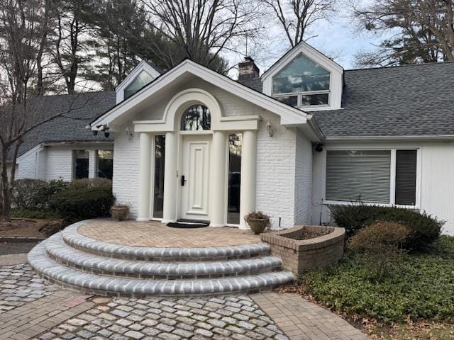 doorway to property with brick siding, a chimney, and roof with shingles