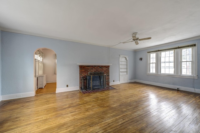 unfurnished living room featuring arched walkways, ceiling fan, baseboards, a brick fireplace, and wood-type flooring