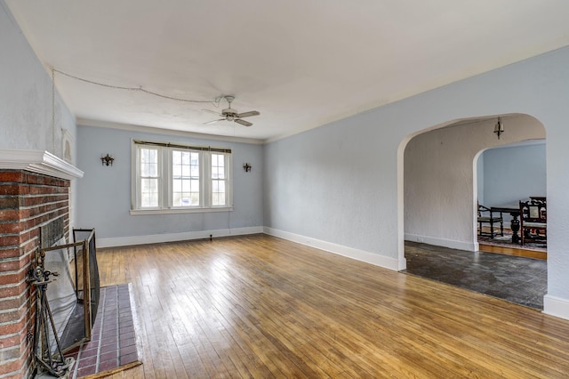 unfurnished living room featuring baseboards, arched walkways, a ceiling fan, hardwood / wood-style floors, and a brick fireplace