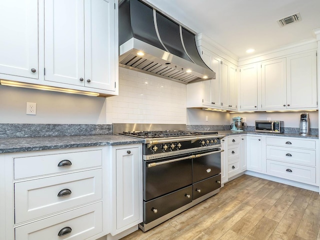 kitchen with range with two ovens, light wood finished floors, visible vents, white cabinets, and wall chimney range hood