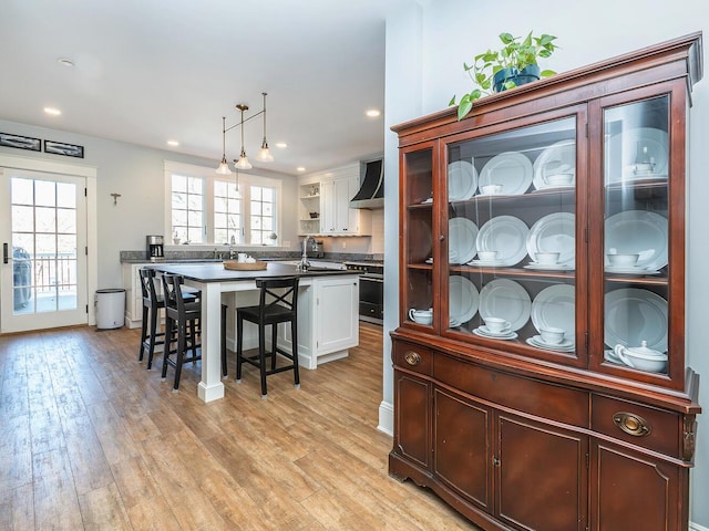 kitchen with dark countertops, wall chimney exhaust hood, light wood-style flooring, a breakfast bar, and white cabinetry