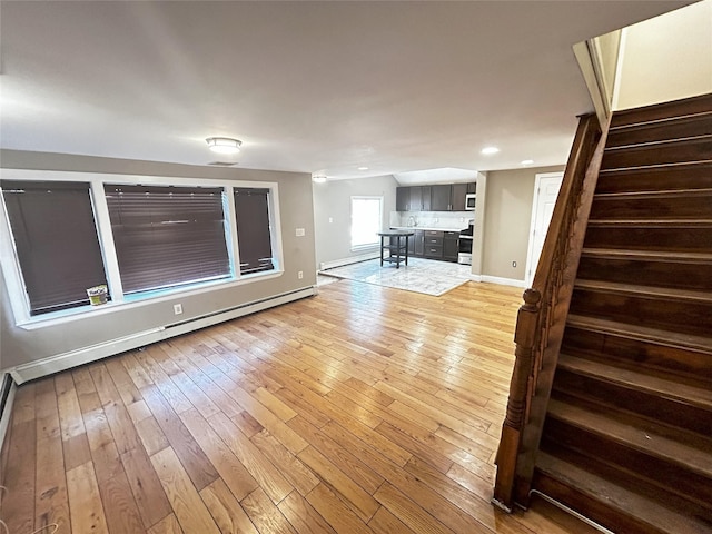 unfurnished living room featuring a baseboard radiator, baseboards, light wood finished floors, and stairs