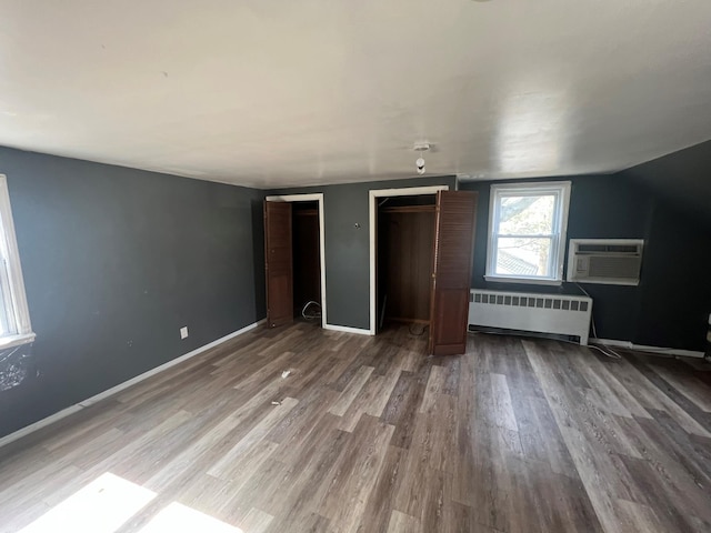 unfurnished bedroom with a wall unit AC, two closets, radiator, wood finished floors, and baseboards