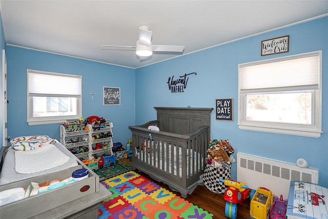 bedroom featuring a nursery area, ceiling fan, wood finished floors, and radiator