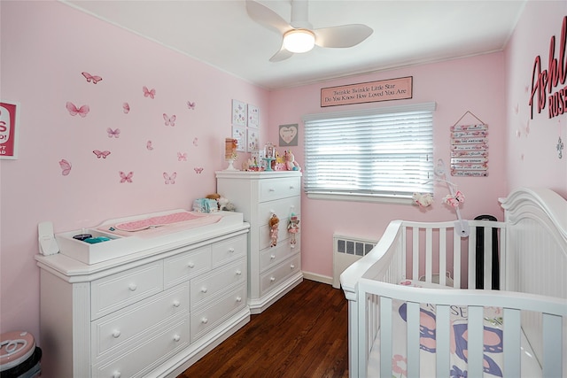 bedroom with dark wood finished floors, radiator heating unit, ceiling fan, a crib, and baseboards